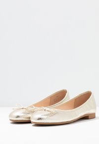 Guldiga ballerinaskor med en slät metallisk finish, rund tå och en liten rosett på framsidan. Platt sula för enkel användning.