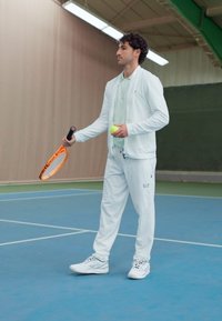 Tenue de sport blanche avec un t-shirt vert clair, tenant une raquette de tennis et une balle, debout sur un court de tennis bleu.