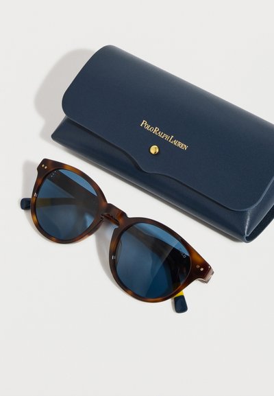 Runde solbriller i skilpaddemønster med mørkeblå linser ved siden av et marineblått Polo Ralph Lauren-etui med gullknapp og logo.