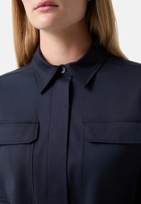 Navy button-up shirt with a fold-down collar, two front pockets, and a smooth texture. Features contrast stitching along the seams.