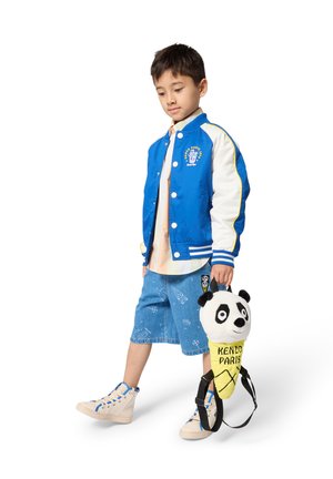Veste bomber bleue à manches blanches, chemise claire, short en denim avec motifs, et un sac à dos en forme de panda. Baskets crème avec des accents bleus.