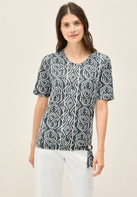 Top de manga corta en azul oscuro con patrón geométrico blanco; escote redondo; detalle anudado en el lateral; tejido ligero y texturizado.