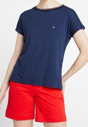 Person trägt ein marineblaues kurzärmeliges T-Shirt mit kleinem Logo und leuchtend roten, knielangen Shorts und steht mit einer Hand in der Hüfte.
