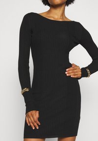 Czarna prążkowana sukienka z długimi rękawami, dekoltem w łódkę i dopasowanym krojem. Ozdobiona złotymi łańcuszkami na nadgarstkach.