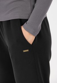 Grijze geribbelde lange mouw top met een gebogen zoom, gecombineerd met zwarte joggers met een gouden accent nabij de zak, gladde stofstructuur.