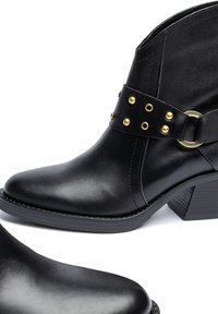Zwarte leren enkellaarsjes met een puntige neus, voorzien van een met goudbeslagen riem en ronde gespen, geplaatst op een gestapelde hak.