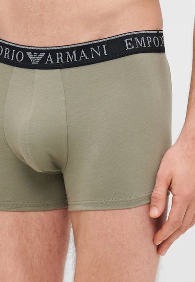 Tamsiai žali medvilniniai apatiniai šortai su juodu, prekės ženklo juosmeniu, kuriame užrašyta "Emporio Armani" metalizuotu šriftu, lygia tekstūra ir prigludusia forma.