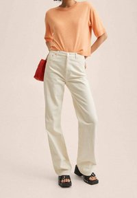 Top in maglia a maniche corte color pesca, jeans bianchi a vita alta e gamba dritta, sandali neri con plateau e una piccola borsa rossa texturizzata.