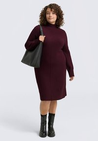 Bordo pletena obleka z visokim ovratnikom in dolgimi rokavi, s pravo silhueto. Oblečena z belo črno torbo in črnimi gležnarji.