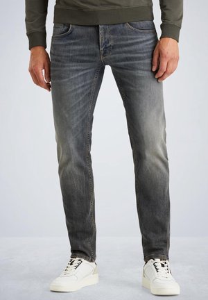 Mężczyzna w wyblakłych szarych dopasowanych dżinsach i białych trampkach, stojący z rękami swobodnie opartymi wzdłuż ciała na jednolitym tle.