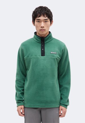 Jeune homme portant un pull polaire vert Columbia avec des boutons-pression noirs et un pantalon gris, face à un fond uni.