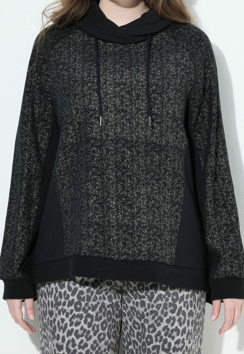Personne portant un sweat à capuche noir et gris tacheté avec des cordons et un pantalon gris à imprimé léopard sur un fond uni.