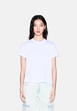 Giovane donna con capelli lunghi neri che indossa una T-shirt bianca Armani Exchange e pantaloni cargo azzurri contro uno sfondo bianco uniforme.