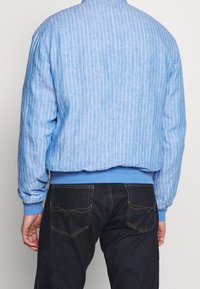 Person som bär en ljusblå randig jacka och mörkblå jeans, vänd bort från kameran mot en enfärgad bakgrund.