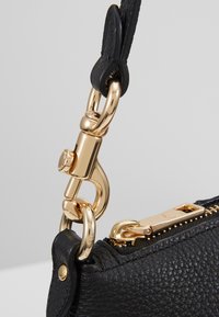 Borsa in pelle nera con superficie testurizzata, dotata di hardware dorato e chiusura con cerniera. Clip e dettagli sono visibili sulla tracolla.