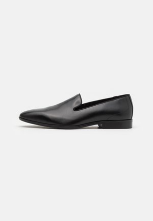 Mocasín masculino de cuero negro con tacón bajo, punta puntiaguda y diseño sin cordones, mostrado de perfil sobre fondo blanco.