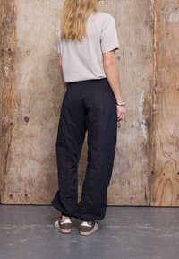 Pantalon large noir texturé, taille élastique, associé à un t-shirt beige court et des baskets à rayures marron.