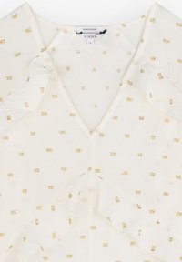 Blouse blanche avec un col en V, ornée de petits motifs carrés dorés, fabriquée en matériau léger, avec des détails de tissu fluide au niveau des épaules.