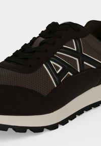 Baskets athlétiques noires et grises avec des lacets noirs, tissu en mesh, logo noir en relief "AX" sur le côté, et une semelle intermédiaire blanche avec une semelle extérieure noire texturée.