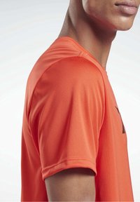 Camiseta deportiva naranja de manga corta, fabricada en un tejido suave con un ligero brillo, que presenta un diseño gráfico negro en la parte frontal.