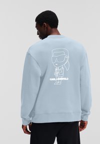 Svetlo modra majica s kapuco z belo obrobno ilustracijo lika v sončnih očalih in suknjiču na hrbtni strani, z teksturirano površino.