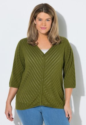 Femme aux cheveux châtain clair mi-longs, portant un cardigan vert à col en V boutonné avec des rayures diagonales et un jean bleu, debout contre un mur blanc.