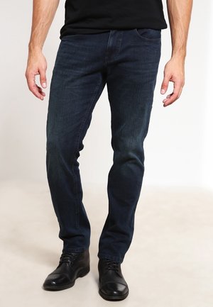 Homme portant un jean bleu foncé et des chaussures en cuir noir, debout devant un fond clair uni.