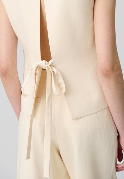 Haut sans manches beige avec une découpe en forme de clé dans le dos et un noeud de fermeture, présentant une texture lisse et une coupe ajustée, associé à un pantalon assorti.