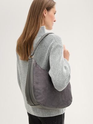 Femme aux longs cheveux bruns portant un pull gris rayé, tenant un grand sac d'épaule en suède et cuir gris foncé sur son épaule droite.