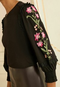 Fekete blúz, rózsaszín és zöld virágos hímzéssel az ujjakon, fodros részletekkel és ráncolt mandzsettával. Bőr szoknyával viselve.