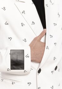 Ręka dostosowująca biały żakiet z czarnymi abstrakcyjnymi symbolami i czarną skórzaną łatą na mankiecie z etykietą "PILAWSKI".