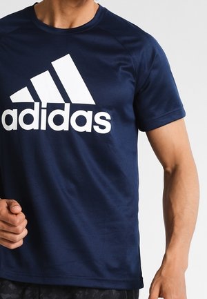 Mężczyzna w granatowej koszulce Adidas z dużym białym logo Adidas na piersi, stojący na jednolitym tle.