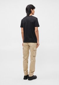 Melnas kokvilnas T-krekls saskaņots ar bēšām biksēm ar sānu kabatām. Apģērbs pabeigts ar melnu apavu. Parādīts skats no aizmugures.