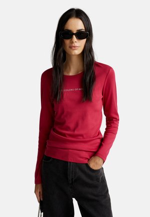 Mujer con cabello largo y oscuro, usando gafas de sol negras, una camisa de manga larga roja y pantalones negros, con una mano en el bolsillo.