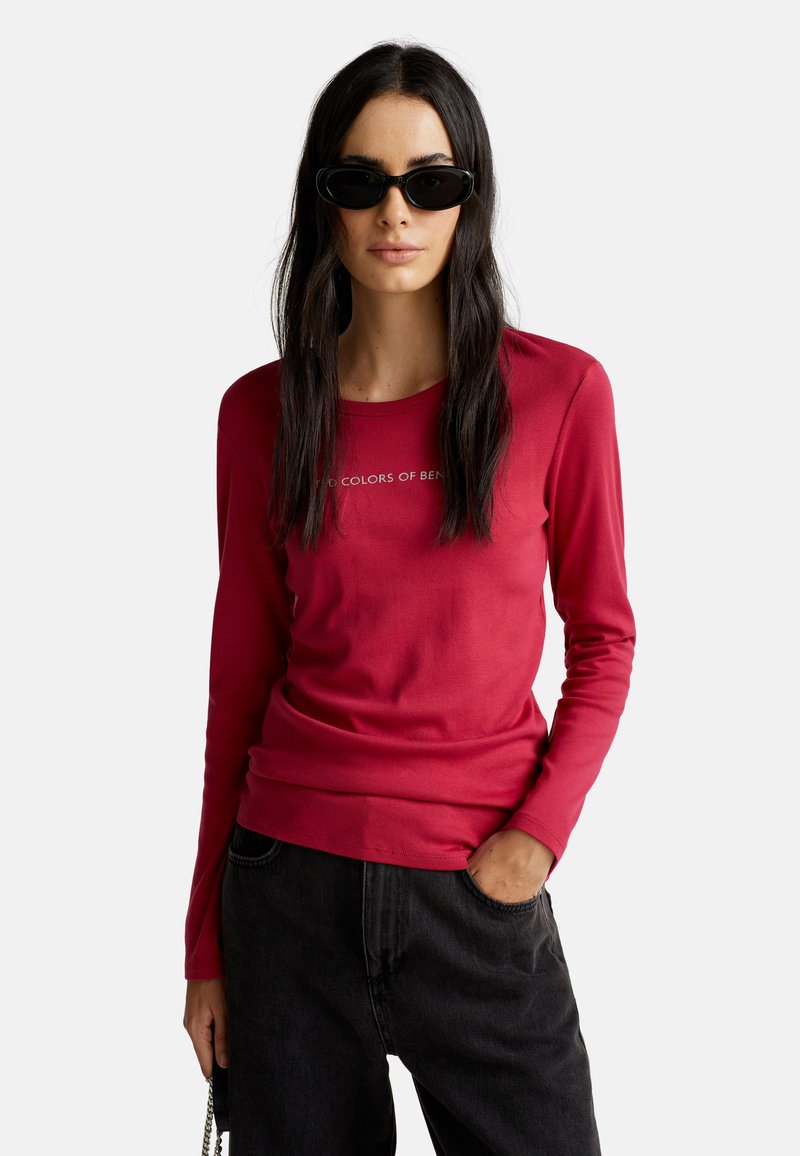 Frau mit langen, dunklen Haaren, die schwarze Sonnenbrillen trägt, ein rotes Langarmshirt und schwarze Hosen hat, mit einer Hand in der Tasche.