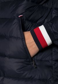 Granatowa pikowana kurtka puchowa w zbliżeniu, pokazująca kieszeń na zamek i mankiet w paski w kolorach czerwonym, białym i granatowym. Gładka, pikowana faktura.