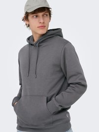 Jonge man met een grijze hoodie met voorzak en trekkoorden, en een lichtbeige pet, die naar links kijkt.