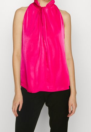 Femme portant un chemisier sans manches rose vif avec un col haut noué et un pantalon noir, montrée des épaules aux hanches.