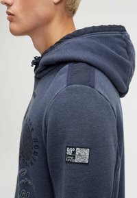 Navy blue hoodie with a textured finish, featuring a drawstring hood, subtle logo print, and a fabric patch on the left sleeve.