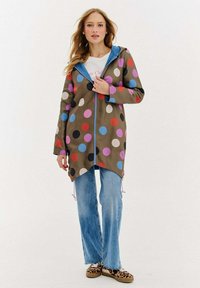 Kobieta w długim oliwkowym płaszczu w wielokolorowe kropki, jasnoniebieskich dżinsach i butach w panterkę, stojąca na białym tle.