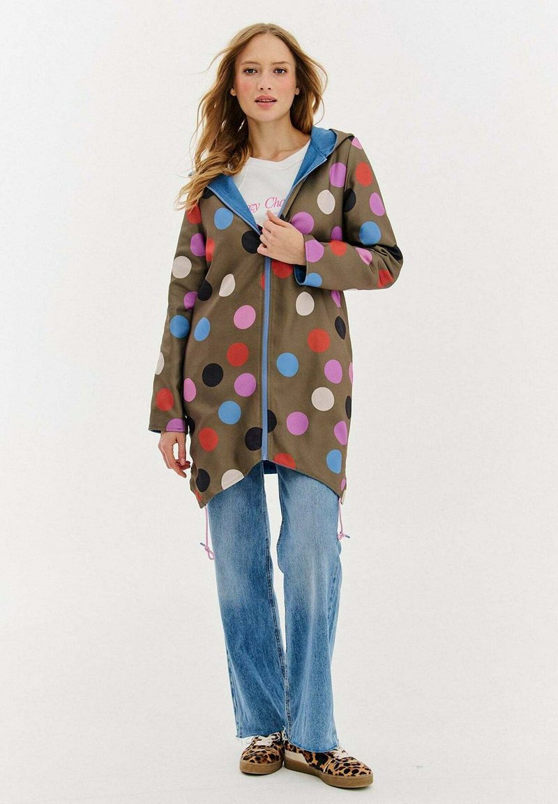 Kobieta w długim oliwkowym płaszczu w wielokolorowe kropki, jasnoniebieskich dżinsach i butach w panterkę, stojąca na białym tle.