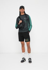 Športový outfit pozostávajúci z trička s dlhým rukávom v zeleno-čiernom gradientnom prevedení, čiernych šortkách s nápisom "Rivonia", tenisiek a čiernej crossbody tašky.
