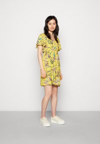 Vestido amarelo com flores, com mangas curtas e franzidas e uma cintura ajustada, apresentando um padrão botânico colorido. Usado com ténis brancos de plataforma.