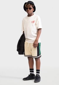 Crème t-shirt met rode graphic, beige shorts met groene en zwarte accenten, zwarte bucket hat, zonnebril en zwarte sneakers met gestreepte sokken.