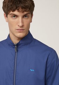 Marinblå zip-up jacka med ståkrage, med en ljusblå broderad hundlogotyp på vänster bröst, slät tygtextur.