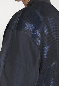 Närbild av en mans axel som visar ett texturerat svart tyg med ett blått kamouflagemönster och en slät svart sektion, vilket indikerar en stilren jacka.