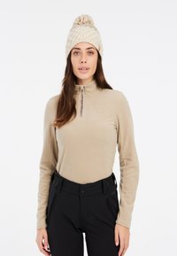 Pullover in pile beige con colletto a mezza zip, abbinato a pantaloni neri a vita alta. Il modello indossa un berretto lavorato a maglia color crema con pom-pom.
