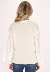 Weißes Hemd mit langen, ausladenden Ärmeln und diamantenförmigen Ausschnitten. Hergestellt aus leichtem Stoff mit glatter Textur.