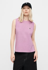 Γυναίκα με ξανθά μαλλιά μέχρι τους ώμους, που φοράει μαύρο καπέλο, μωβ αμάνικη μπλούζα με μικρό λογότυπο και μαύρο παντελόνι, μπροστά σε απλό φόντο.