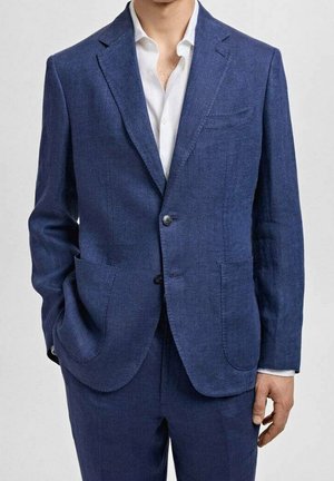 Mann trägt einen blauen Zweiknopf-Blazer und passende Hose mit weißem Hemd, linke Hand in der Tasche, vor einfarbigem Hintergrund.